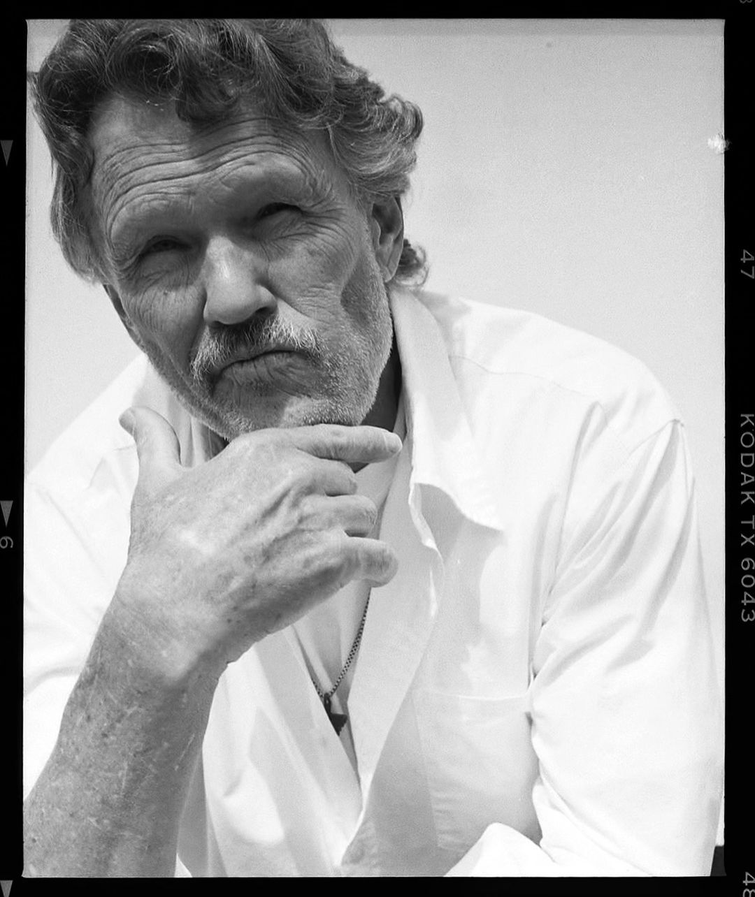 Black and white portrait of a mature man with a mustache wearing a white shirt and resting his chin on his fingers.