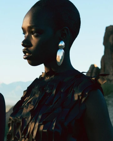 Black woman wearing large silver drop earrings and a textured black top against a rocky outdoor background.