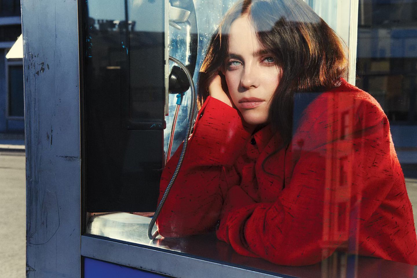 Portrait of a woman in a red jacket inside a phone booth with an urban background, shot in medium close-up with natural lighting.