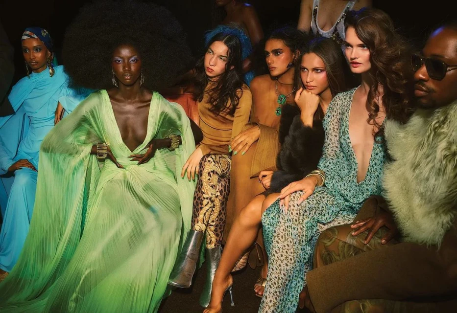 Diverse group of models in a fashion show audience wearing different outfits, including dresses, furs, and glasses in a dimly lit room.