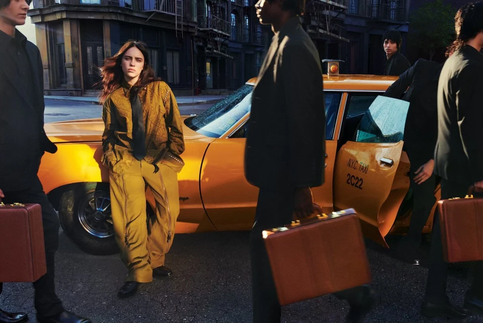 Fashion editorial shot of a female leaning against a yellow taxi cab in an urban setting, surrounded by people holding briefcases.