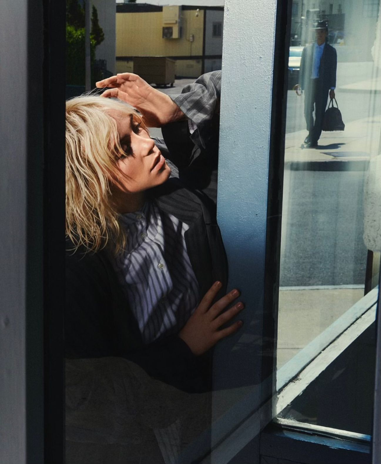 Woman with blonde hair poses in a gray suit reflected in a city window with a man in a suit walking by in the background.