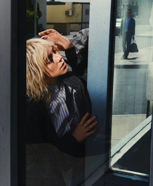 Woman with blonde hair poses in a gray suit reflected in a city window with a man in a suit walking by in the background.