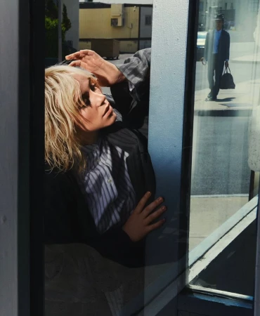 Woman with blonde hair poses in a gray suit reflected in a city window with a man in a suit walking by in the background.