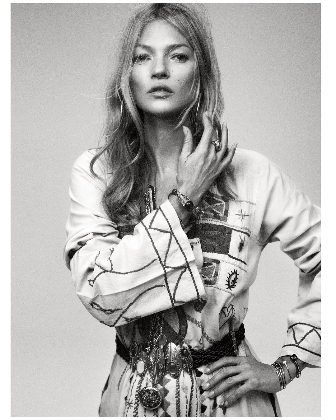 Monochrome portrait of a fashionable adult woman with long blond hair, wearing an embroidered tunic and multiple pieces of jewelry.