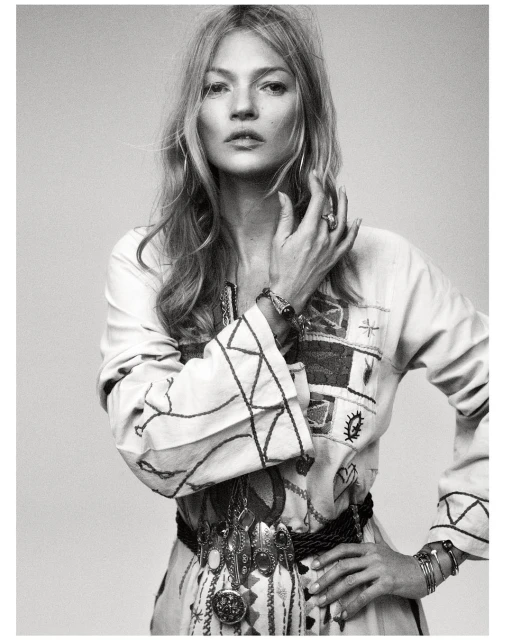 Monochrome portrait of a fashionable adult woman with long blond hair, wearing an embroidered tunic and multiple pieces of jewelry.