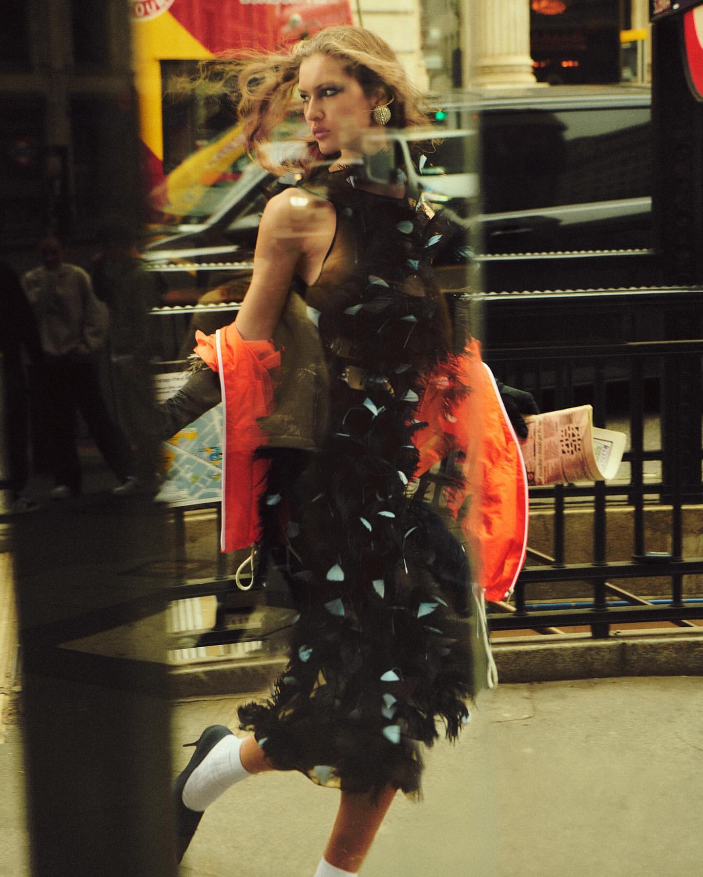 Fashionable woman strides through city street wearing a feather boa, orange jacket, and heels, reflected in a passing vehicle.