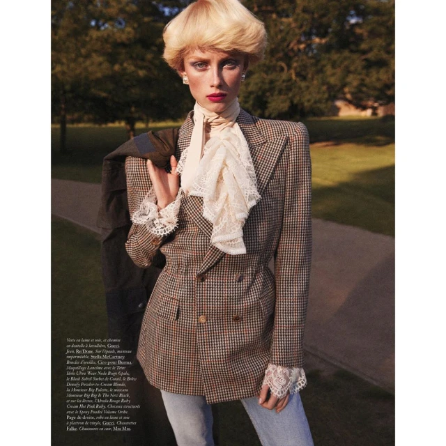 Editorial fashion shot of a blonde model in a brown plaid blazer with a ruffled collar posing in a park with green trees.