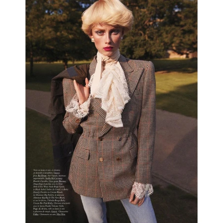 Editorial fashion shot of a blonde model in a brown plaid blazer with a ruffled collar posing in a park with green trees.
