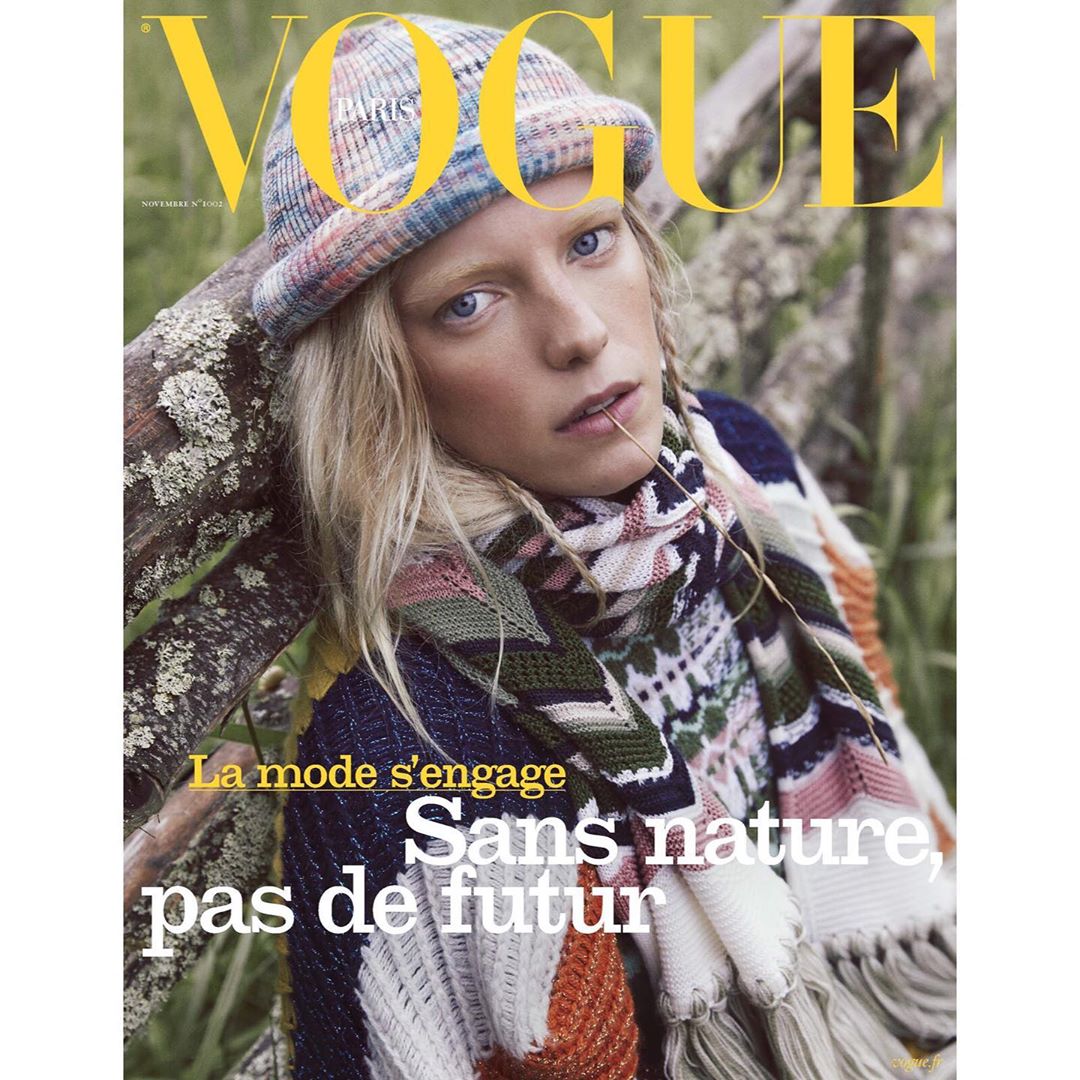 Vogue cover featuring a blonde woman in a knit hat and patterned scarf, leaning against a mossy branch with the magazine's logo.