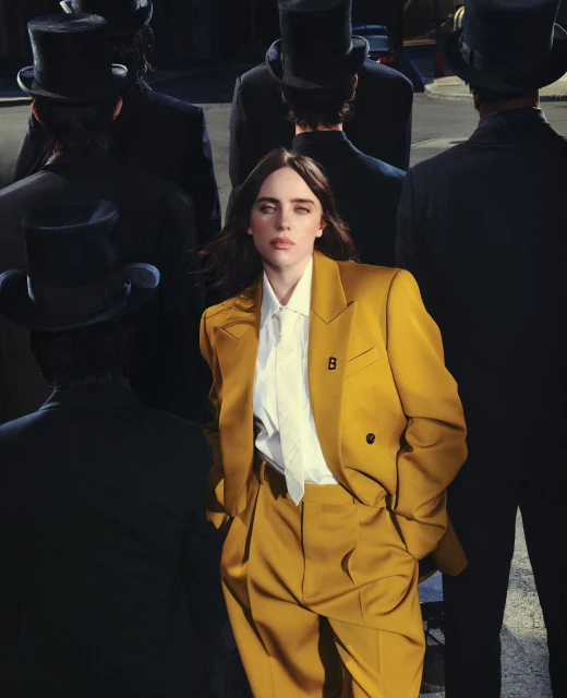 Singer Billie Eilish in a mustard-yellow suit, surrounded by men in dark suits and top hats, creates a striking fashion editorial image.