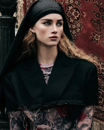 Woman in black jacket and headscarf poses in front of a patterned rug, creating an editorial fashion portrait with soft, diffused lighting.