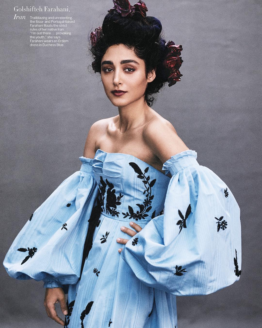 Elegant woman in a light blue off-the-shoulder dress with black floral embroidery and elaborate hairstyle adorned with flowers, portrait style.