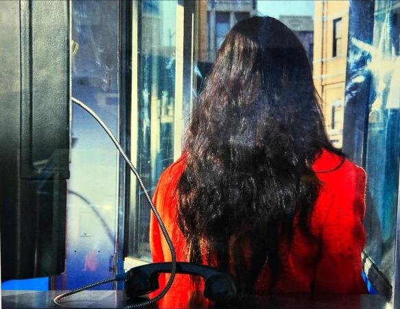 Woman in red jacket inside a public payphone booth in urban environment, city buildings are visible through the glass.