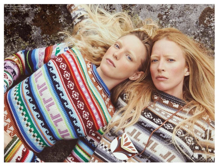 Two fair-skinned adult women with long blond hair lie next to each other wearing patterned knit sweaters against a rocky surface.