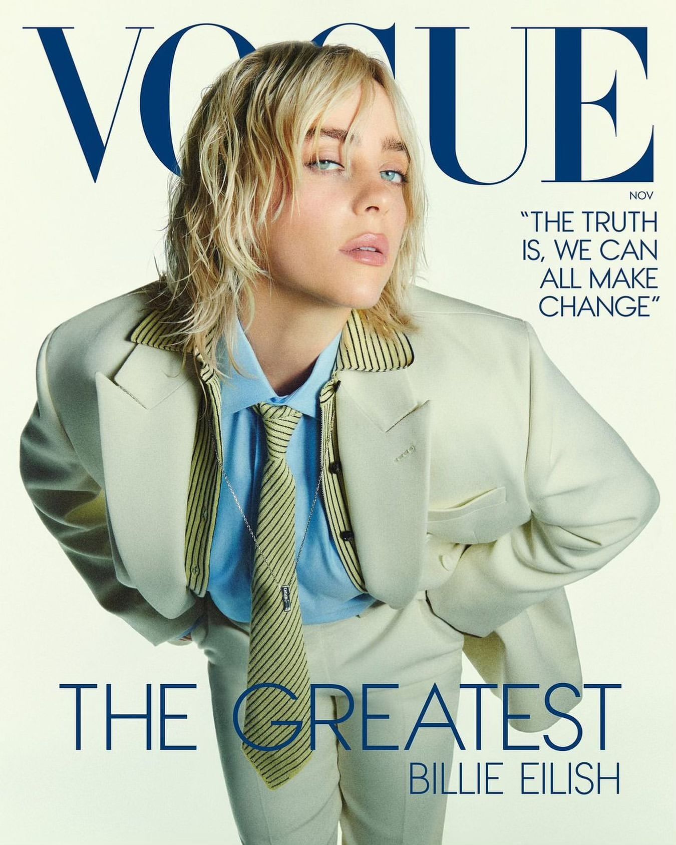 Billie Eilish, the cover star of Vogue, poses in a cream-colored suit and striped tie in a studio setting, her blonde hair tousled.