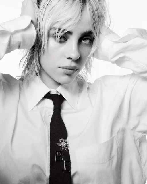 Black-and-white portrait of Billie Eilish wearing a white collared shirt and black tie with her hands behind her head.