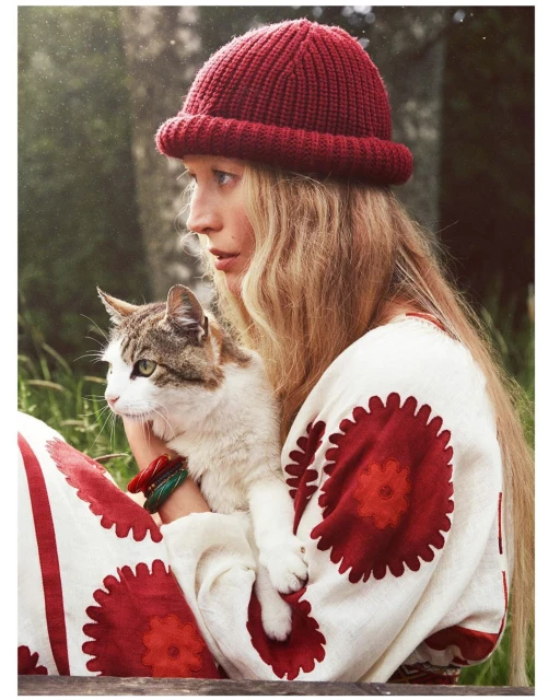 Woman with long blonde hair and a red knit hat holds a tabby cat in her arms, posing outdoors with a blurred green background.