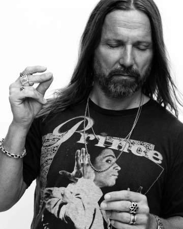Black and white portrait of a man with long hair wearing a graphic t-shirt and multiple rings by Mikael Jansson