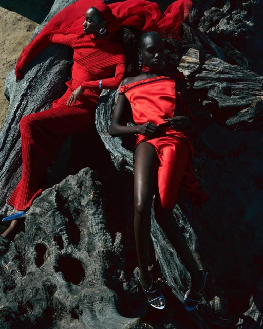 Two women in red outfits pose on a textured tree trunk in a sunlit outdoor setting, showcasing fashion and style.