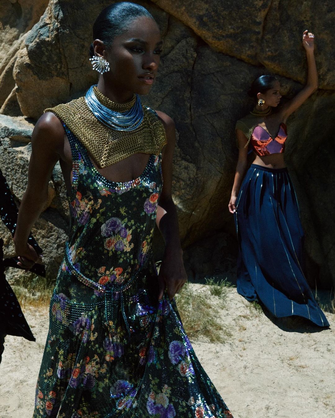 Two women pose among rocks, one wearing a floral dress with metallic accents and the other in a crop top and long skirt.
