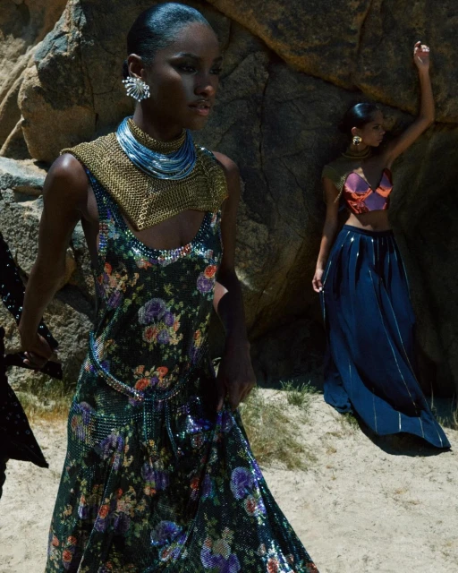 Two women pose among rocks, one wearing a floral dress with metallic accents and the other in a crop top and long skirt.