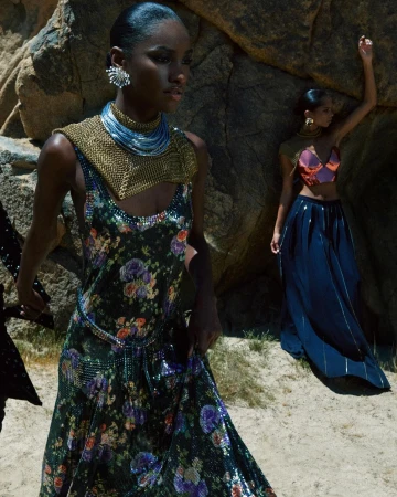 Two women pose among rocks, one wearing a floral dress with metallic accents and the other in a crop top and long skirt.