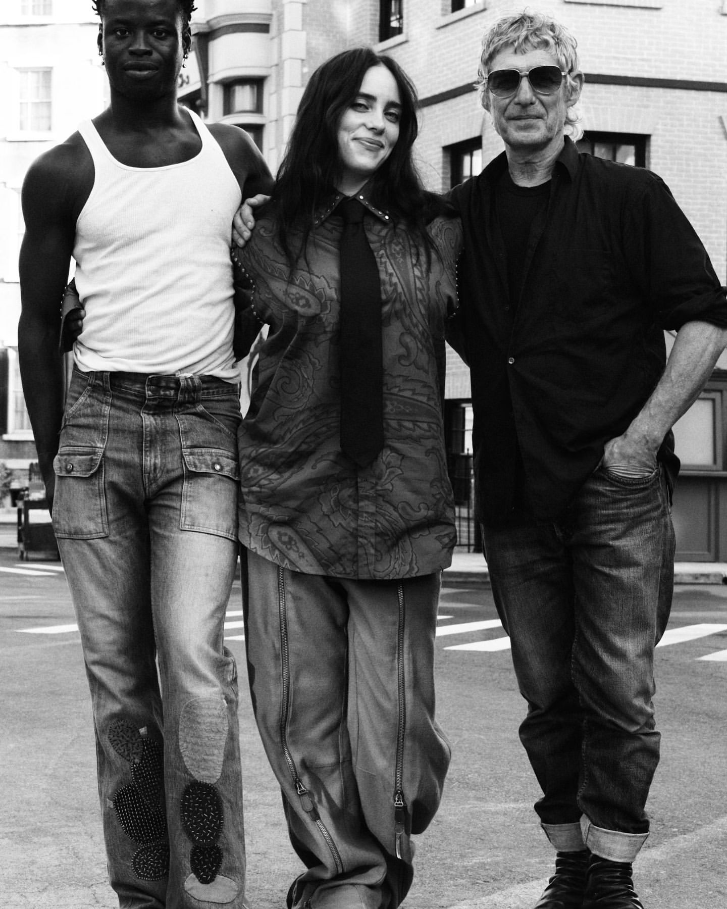 Three people stand close together: two men, one with dark skin in a white tank top, a woman in a patterned top and tie, city background.