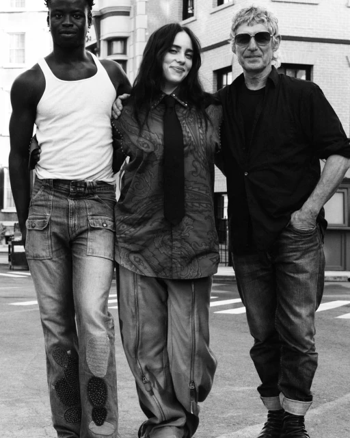 Three people stand close together: two men, one with dark skin in a white tank top, a woman in a patterned top and tie, city background.