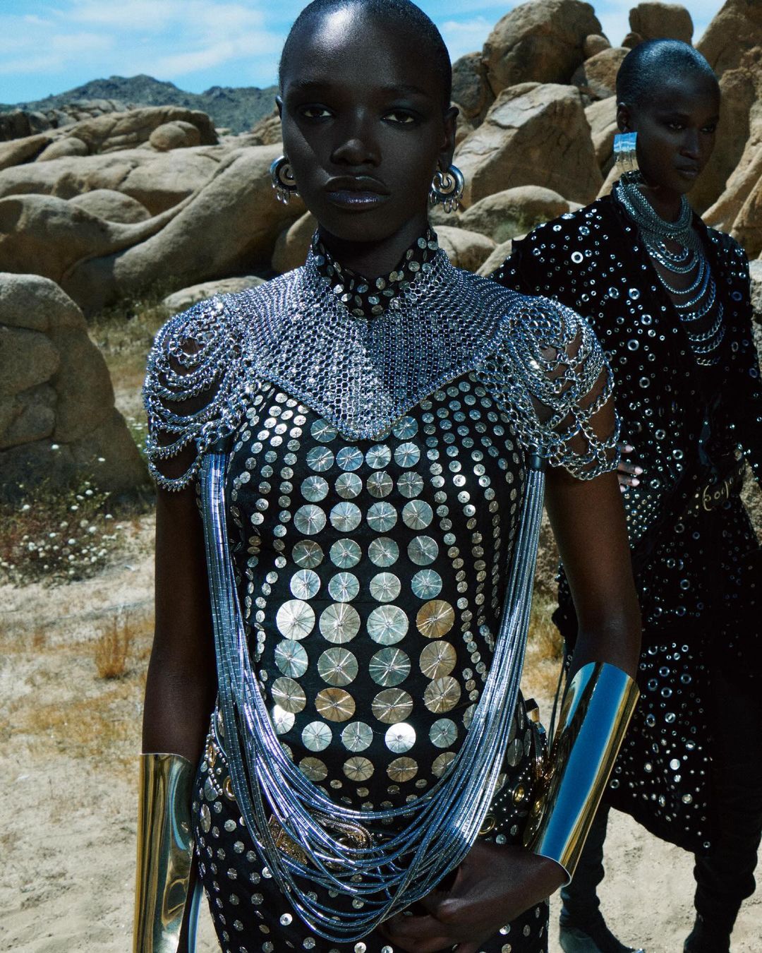 Two African American women posing in a desert landscape, wearing embellished black dresses, statement jewelry, and metallic arm cuffs.