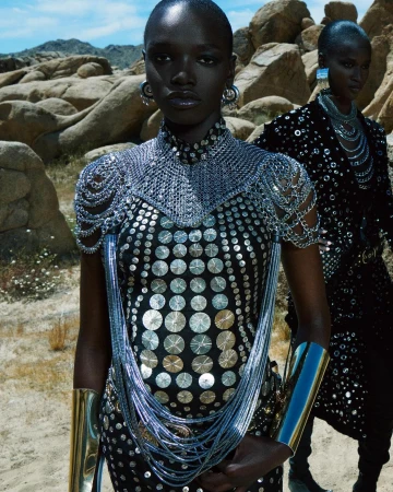 Two African American women posing in a desert landscape, wearing embellished black dresses, statement jewelry, and metallic arm cuffs.