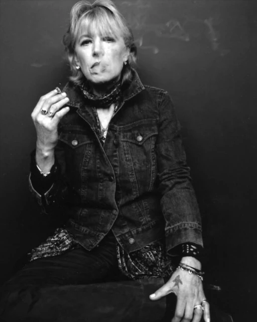 Monochrome portrait of a mature woman smoking, wearing a denim jacket and patterned top against a dark backdrop, exuding a relaxed mood.