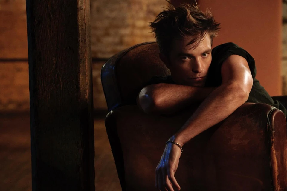 Portrait of a young adult man in a dark t-shirt sitting in a vintage chair in a dimly lit room, resting his arm on the chair.