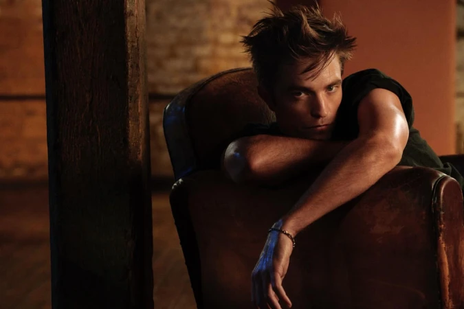 Portrait of a young adult man in a dark t-shirt sitting in a vintage chair in a dimly lit room, resting his arm on the chair.