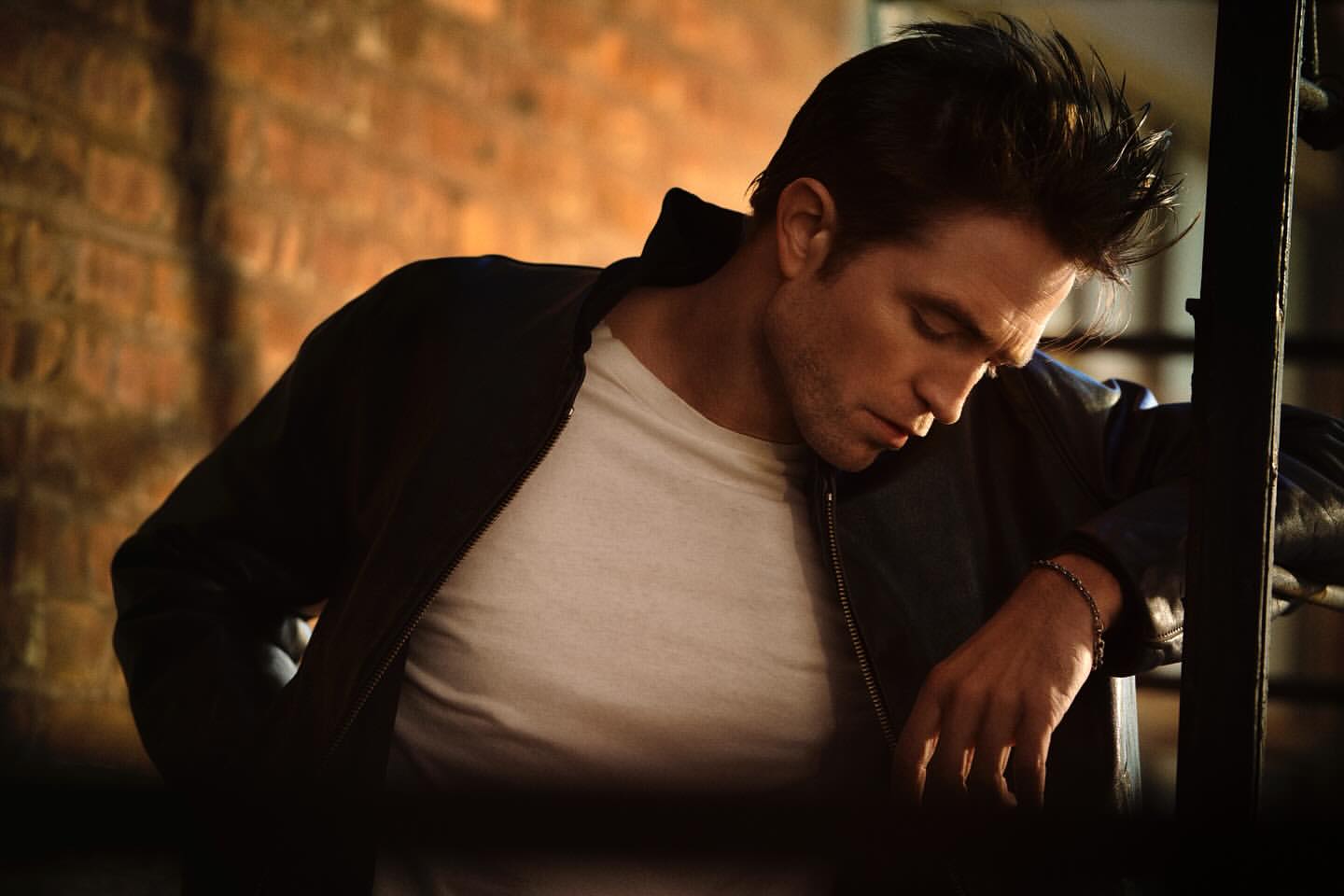 Robert Pattinson in a dark jacket and white t-shirt leans against metal bars in front of a brick wall in a portrait shot.