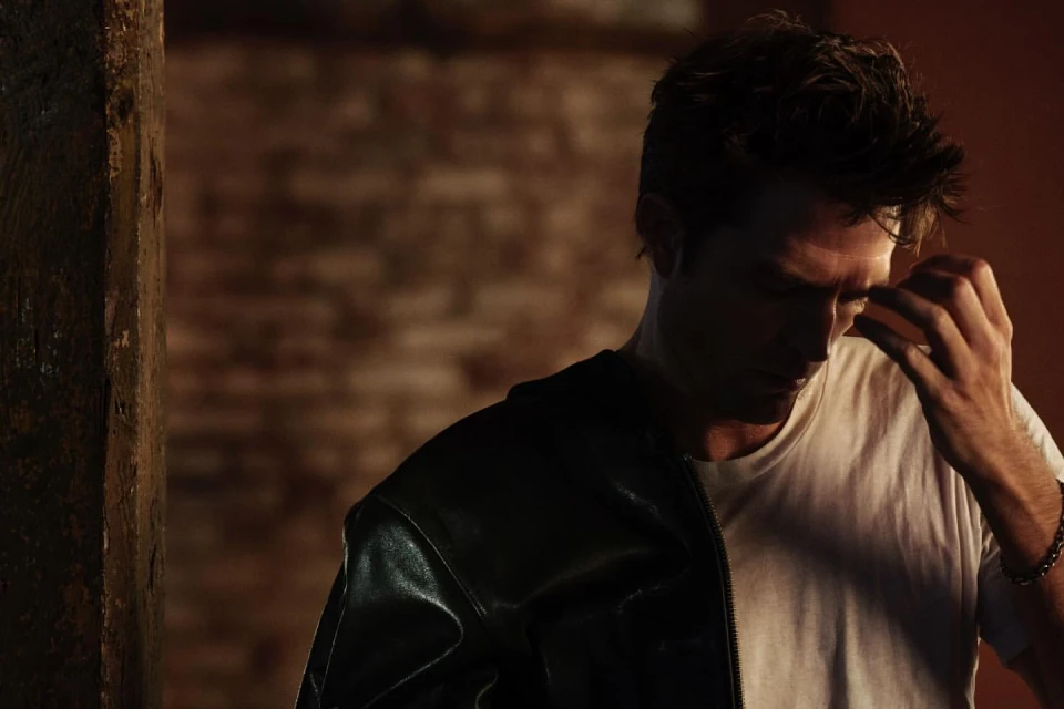 Adult male in white t-shirt and leather jacket in dark lighting against a brick wall, hand raised to face, wrist bracelet visible.