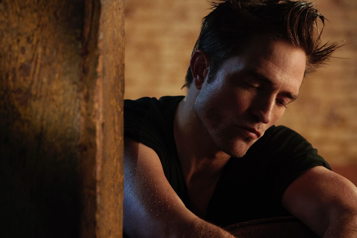 Portrait of a handsome adult male with dark hair, wearing a black shirt, posing indoors with soft lighting near a textured wall.