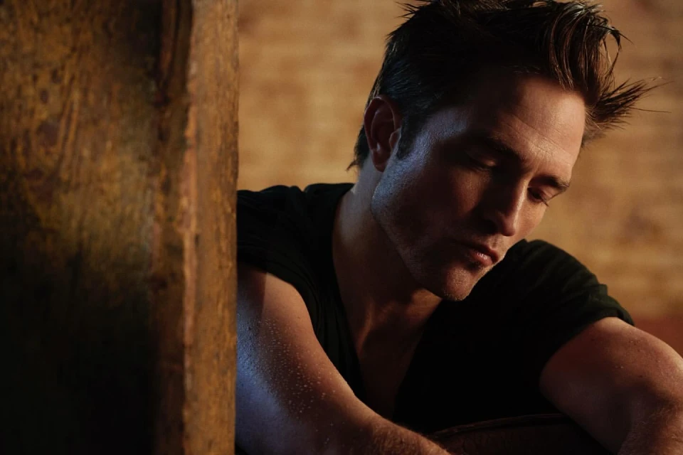 Portrait of a handsome adult male with dark hair, wearing a black shirt, posing indoors with soft lighting near a textured wall.