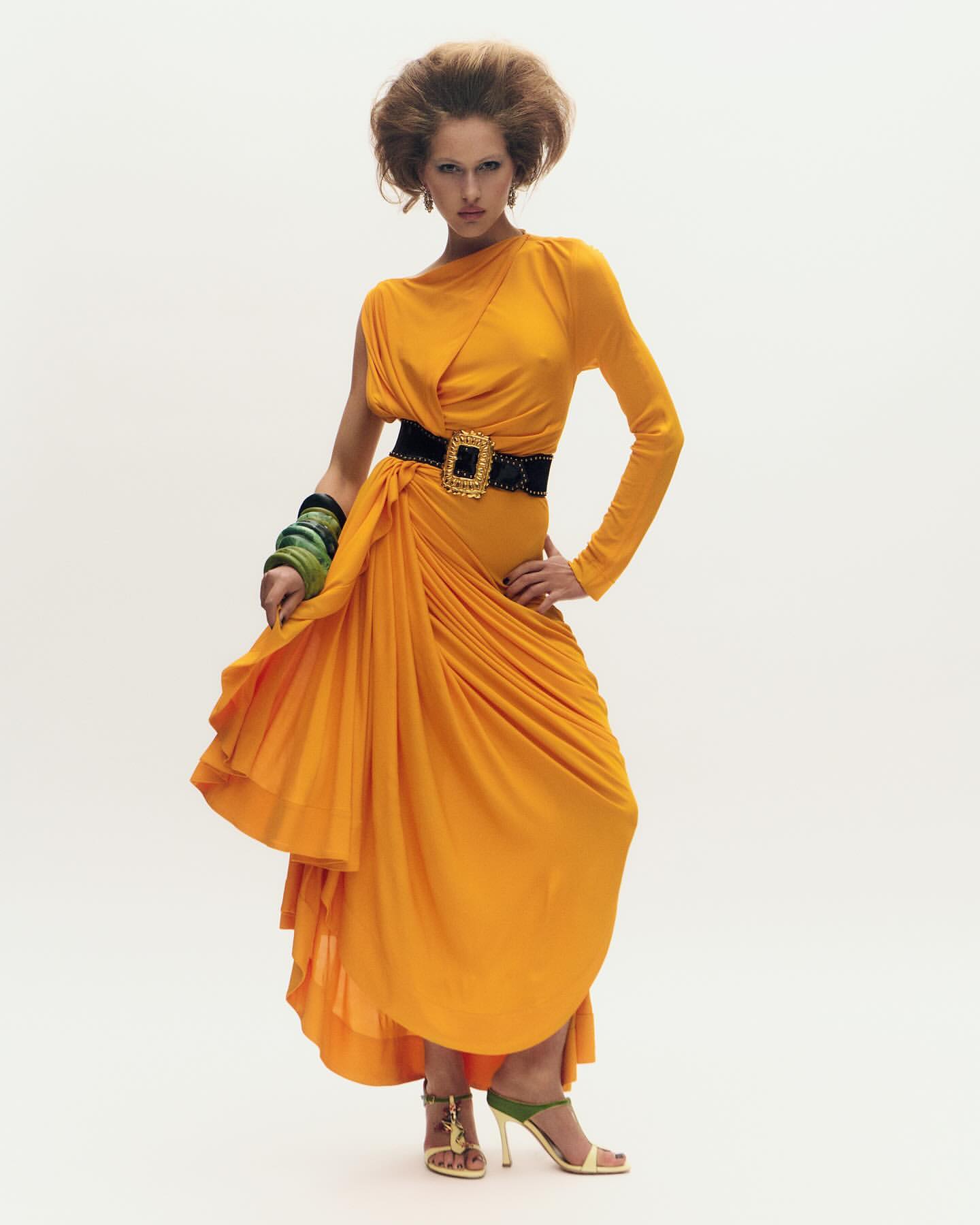 Fashion model poses in a yellow one-shoulder dress with a wide black belt, chunky bracelets, and gold high heels against a white background.