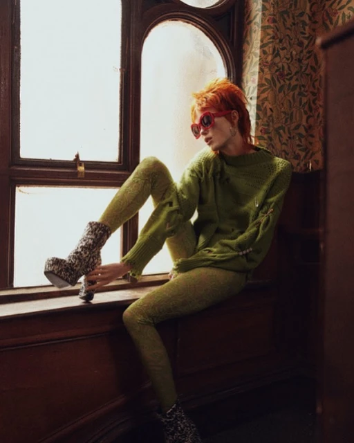 A person with orange hair and sunglasses sits in a window wearing an olive-green sweater, matching leggings, and platform boots with floral pattern.
