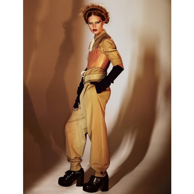 A fashion photo of a woman in tan pants, a tan shirt, and a copper bustier, posing in a photo studio with warm, diffused lighting.
