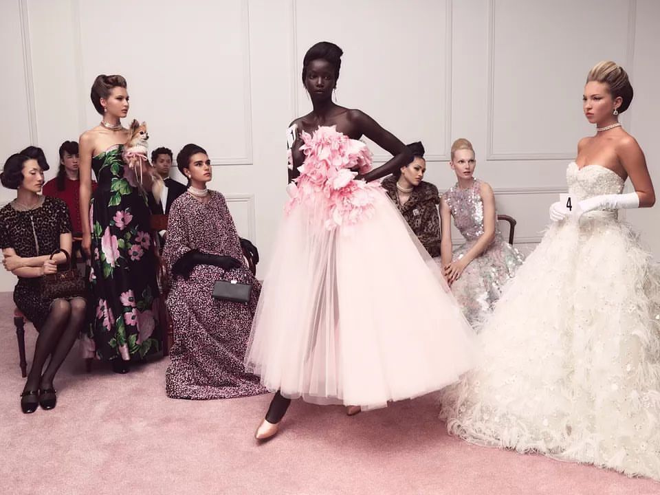 A group of women in various elegant dresses pose in an indoor setting, with a pink carpet and white walls, in a fashion editorial-style photograph.