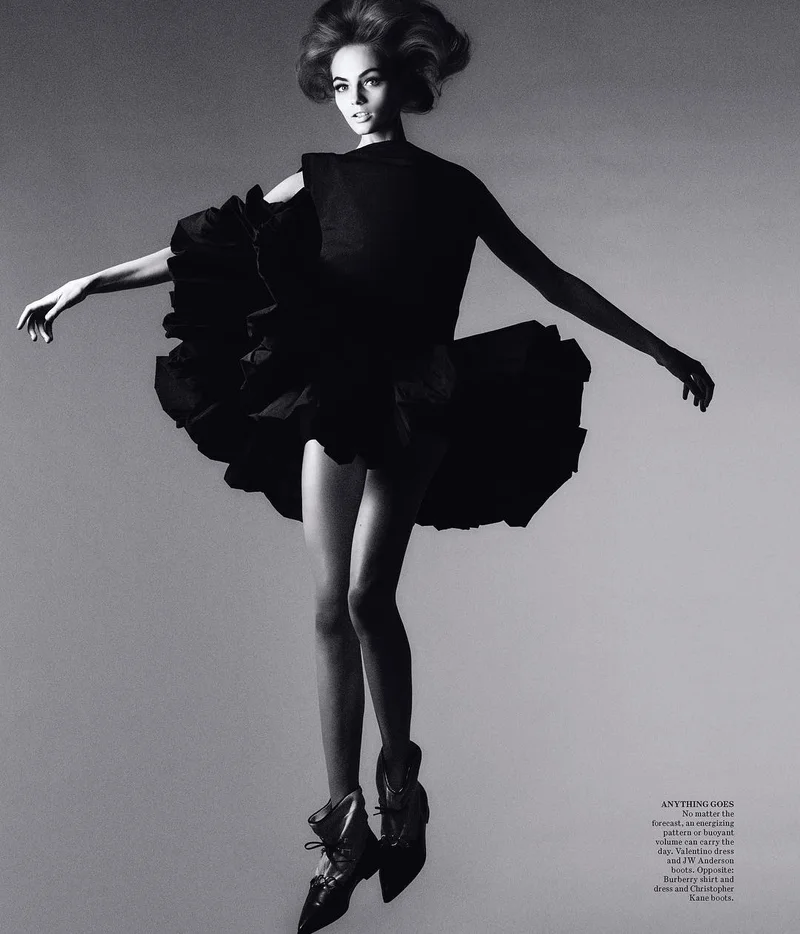 Model in a black ruffled dress with voluminous skirt and boots jumping against a plain grey studio backdrop