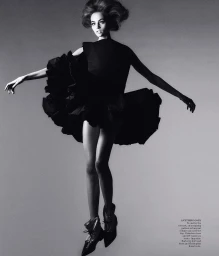 Model in a black ruffled dress with voluminous skirt and boots jumping against a plain grey studio backdrop
