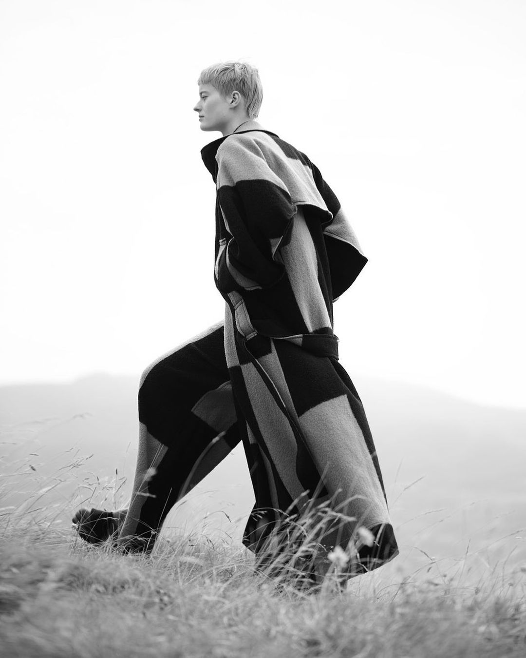 Monochrome shot of a person in a coat and pants walking through a grassy field against a bright sky and distant hills.