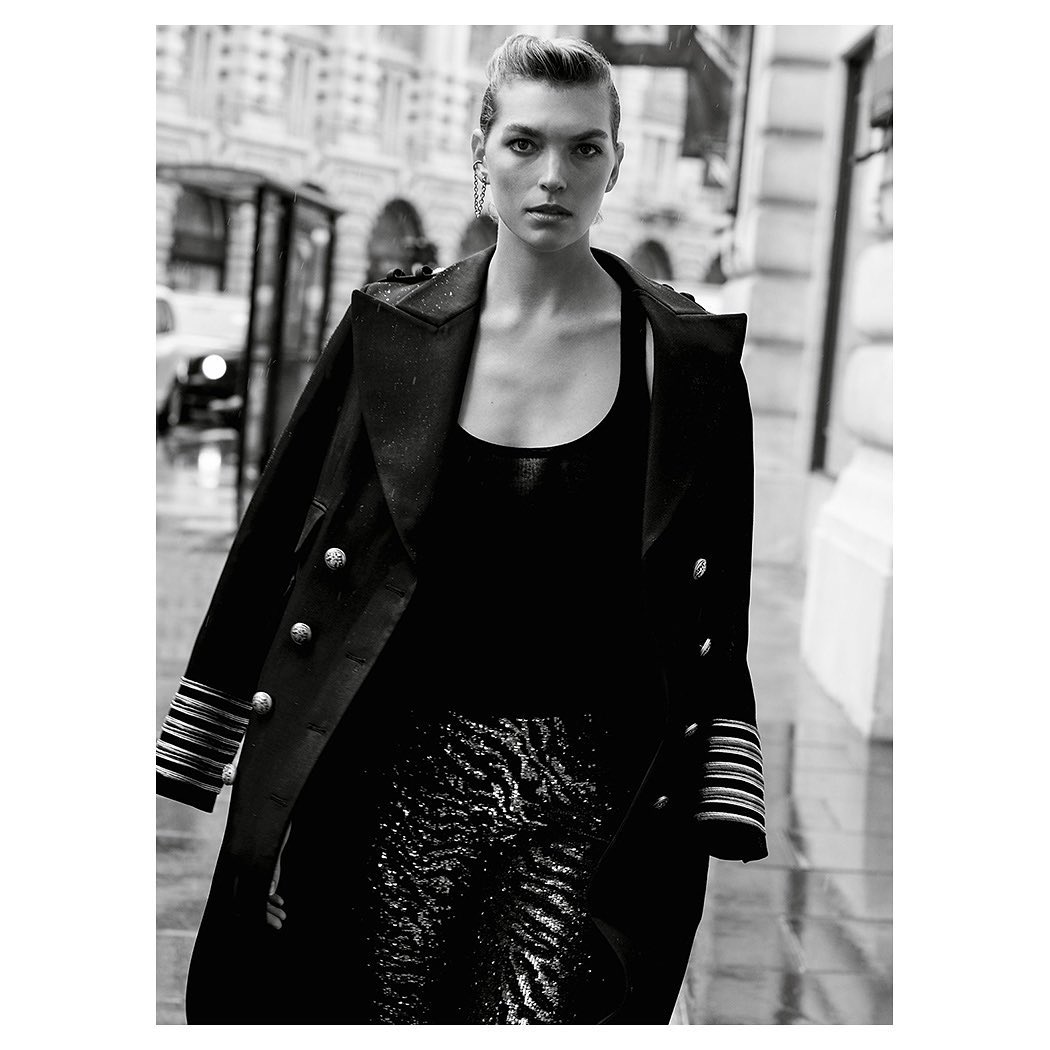 Black and white fashion shot of adult female in long black jacket and patterned pants walking on city street, looking at the camera.