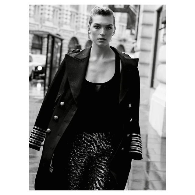 Black and white fashion shot of adult female in long black jacket and patterned pants walking on city street, looking at the camera.