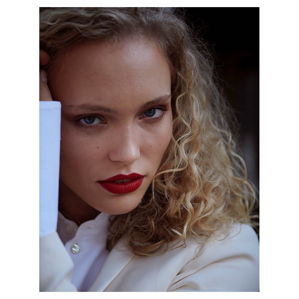 Close-up portrait of a young adult woman with curly blond hair, red lipstick, wearing a white blazer, against a dark background.