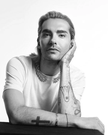 Black and white portrait of a fair-skinned adult male with blonde hair, tattoos, an ornate necklace, and a white t-shirt against a white backdrop.