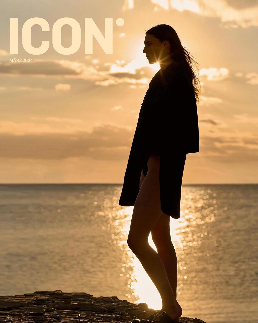 Silhouette of a woman in a long coat standing on a rocky outcrop by the sea at sunset, magazine cover for ICON featuring visible text.
